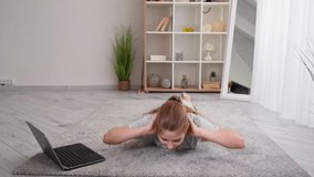Abs sport. Online fitness. Home training. Teenager girl doing workout for back pressing up on floor looking laptop in light room interior.