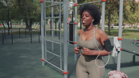 Medium slowmo shot of active young plus size African-American woman in earphones and armband with smartphone running in place having outdoor workout at sportsground