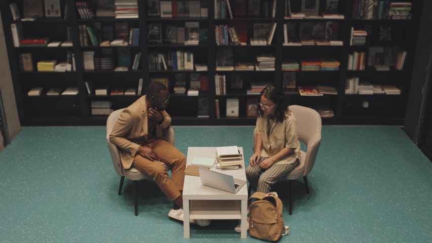 High-angle stab shot of female Asian university or high-school student and young African-American male professor in smart casualwear sitting at table in modern library studying on laptop together