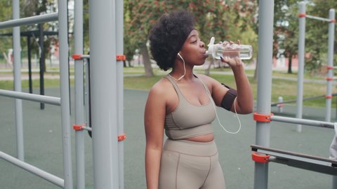 Medium slowmo shot of young plus size African-American woman in tight sportswear drinking water while resting during outdoor workout at sports ground with facilities