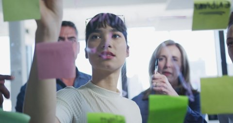 Diverse group of businesswomen and businessman working in a modern office, brainstorming writing on clear board with memo notes, seen through, slow motion