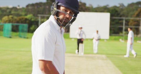 Bircial cricket player on the pitch, turning his head and looking to the camera, before batting during a cricket match, with other players in the background, in slow motion