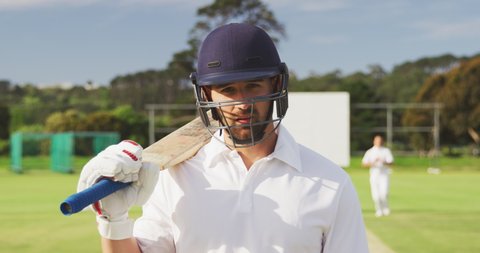 Biracial male cricket player holding a cricket bat on his shoulder, standing on a cricket pitch on a sunny day looking to camera, with other players standing in the background, in slow motion