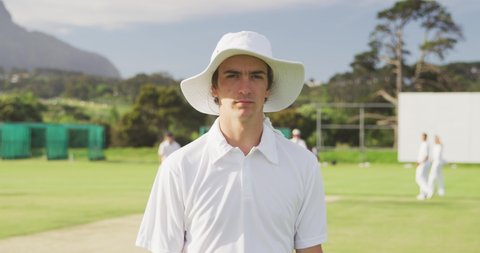 Confident teenage Caucasian male cricket player, standing on a cricket pitch on a sunny day looking to camera, with other players standing  in the background, in slow motion