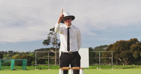 Caucasian male cricket umpire standing on a cricket pitch on a sunny day, signalling to players on the pitch, nodding and pointing his finger in the air, in slow motion