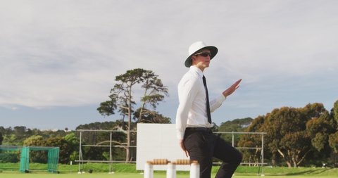 Front view of a Caucasian male cricket umpire standing on a cricket pitch on a sunny day, signalling to players on the pitch, pointing his finger in the air, in slow motion