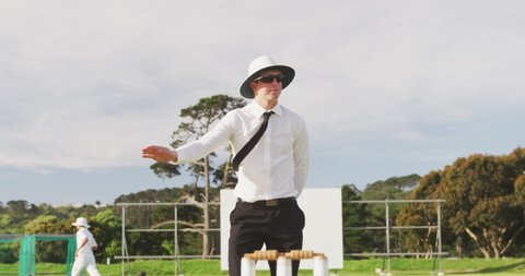 Caucasian male cricket umpire wearing white shirt, black tie, sunglasses and a wide brimmed hat, standing on a cricket pitch on a sunny day, gesturing to players on the pitch in slow motion