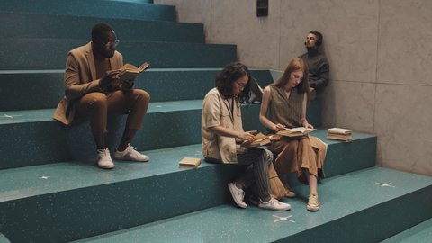 Stab slowmo shot of group of four diverse male and female university students sitting on stairs indoors reading paper books and studying