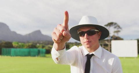 Front view of a Caucasian male cricket umpire standing on a cricket pitch on a sunny day, signalling to players on the pitch, pointing his finger in the air, in slow motion