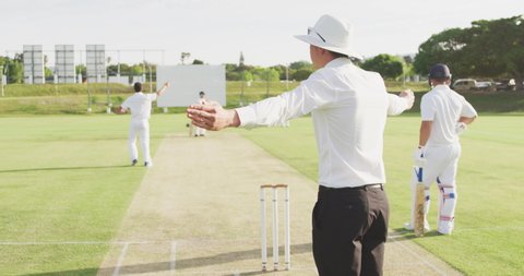 Caucasian male cricket umpire standing on a cricket pitch on a sunny day, signalling to the players on the pitch, gesturing with his hands, with cricket players in the background, in slow motion