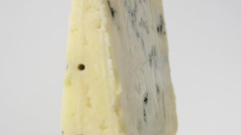 A piece of blue cheese rotates on a white background. Delicacy cheese with green or blue mold, macro view. A dairy product with mold - cheese as a delicacy. Veins of mold in white fresh cheese.