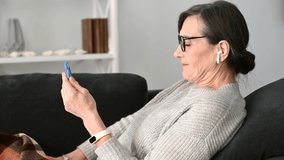 Positive senior mature lady sitting in a relaxed pose on the comfortable couch with a smartphone, mid age woman making a video call, waving hands, greeting friend or family duiring virtual meeting