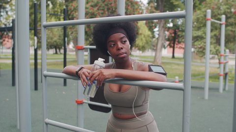 Medium slowmo shot of young African-American woman in sportswear and earphones leaning on sports facility while resting during outdoor training, wearing armband for smartphone