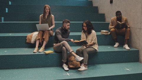 Stab slowmo shot of four young multi-ethnic university students sitting on turquoise stairs indoors reading, studying on laptop and chatting