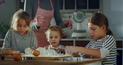 Cinematic authentic shot of happy mother and her little kids are having fun to make dough with flour and knead it together for baking cookies in kitchen at home.