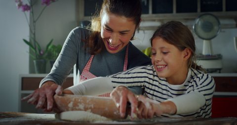 Cinematic authentic shot of happy mother and little daughter are having fun to roll out dough of flour together for baking cookies or bread in kitchen at home.