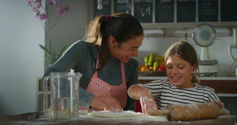 Cinematic authentic shot of happy mother and little daughter are having fun to make dough with flour together for baking homemade cookies or cake in kitchen at home.