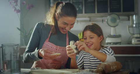 Cinematic authentic shot of happy mother and little daughter are having fun to make forms on dough together for baking homemade cookies in kitchen at home.