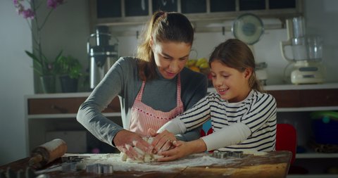 Cinematic authentic shot of happy mother and little daughter are having fun to knead dough with flour together for baking homemade cookies or bread in kitchen at home.