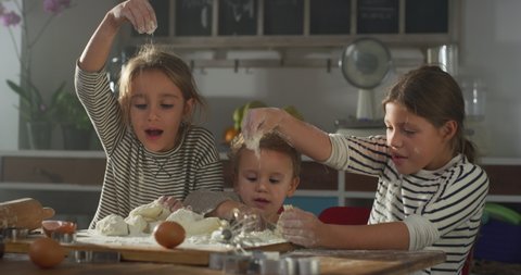 Cinematic authentic shot of happy sisters and little brother are having fun to play together with dough of flour made by their mother for baking cookies in kitchen at home.