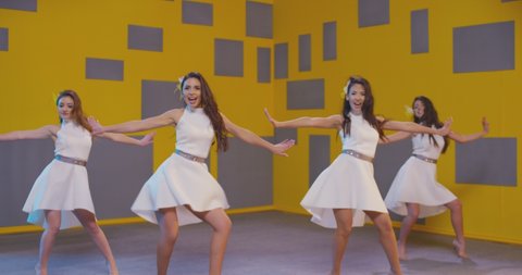 4K Group of beautiful funny stylish girls in white same dresses dancing inside decorated pavilion with grey cubes and yellow wall background . Synchronized dance of female ensemble for video clip ,
