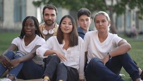 Friendly group of cheerful multinational college students waving hello while sitting on campus lawn. Joyful multiracial classmates smiling while posing for joint portrait video after studies