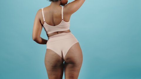 Rear-view medium portrait of curvy young African-American woman in underwear dancing tenderly on isolated blue background. Concept of natural beauty and body positivity