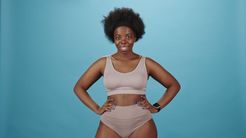 Medium studio portrait of confident young plus size African-American woman in pink underwear holding big white poster with Body Positive lettering standing on blue background