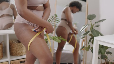 Midsection slowmo shot of young plus size African-American woman in underwear measuring her thighs with yellow measuring tape standing in front of mirror at bedroom