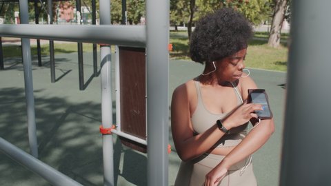 Medium slowmo portrait of smiling young African-American woman in sportswear swiping on smartphone in armband then looking at camera standing outdoors at sports ground with exercise equipment