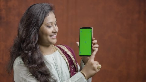 Happy Young indian women using mobile phone by sliding green screen by looking into camera - concept of special offer, discount sale, online shopping and e-commerce