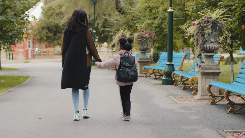 Back view african woman mother holds little afro american girl by hand going in city park back to school study elementary class lesson. Black small pupil with backpack goes home with mum together Royalty-Free Stock Footage #1080931580