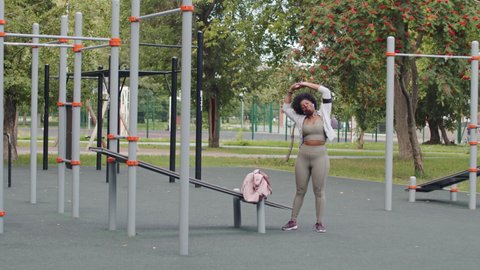 Full-length stab slowmo shot of young plus size African-American woman with handbag on shoulder arriving at outdoor sports ground with exercise facilities for workout