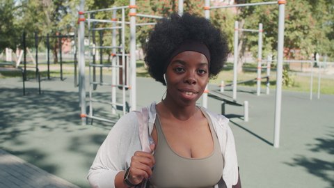 Medium close-up portrait of young active African-American woman posing for camera standing against exercise equipment at outdoor sports ground on sunny day