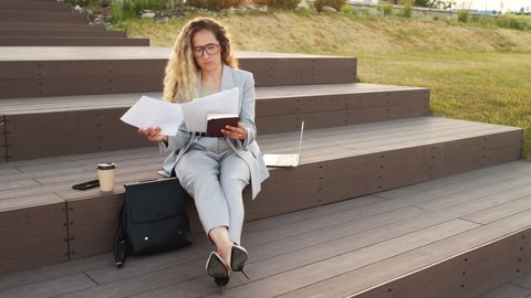 Slowmo stab shot of attractive young businesswoman with long curly blond hair wearing smart casualwear and eyeglasses looking at documents and working on laptop sitting on stairs outdoors at sunset