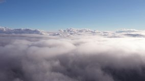 the drone rises above curly clouds with a clear sky and mountains with snowy peaks are visible in the distance
