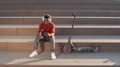 Slowmo stab shot of young cheerful Black man in smart casualwear and headphones sitting on stairs outdoors next to e-scooter watching energetic music video on smartphone, dancing with hands to beat