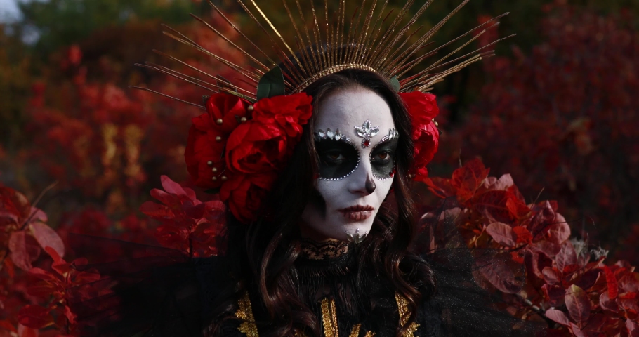 Portrait of young woman with sugar skull makeup and red roses dressed in black costume of death as Santa Muerte against background of autumn forest. Day of the Dead or Halloween concept.