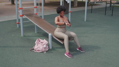 Full-length stab slowmo shot of young plus size African-American woman sitting on gym facility outdoors at sportsground listening to music resting after workout