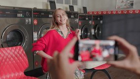 Close up hands hold phone shoots video on background blond woman listens to music dancing on camera sitting in laundry room. Housework service washing machine dryer indoors. Slow motion