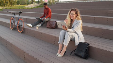 Slowmo stab shot of two young interracial business people sitting on stairs outdoors. Woman having video chat on digital tablet while Black man working on laptop in background
