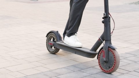 Low-section slowmo shot of unrecognizable male legs in white sneakers standing up on e-scooter and starting ride outdoors