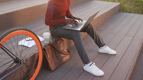 Cropped slowmo shot of unrecognizable African-American businessman or freelancer in smart casualwear sitting on stairs in park next to his bike typing on laptop computer