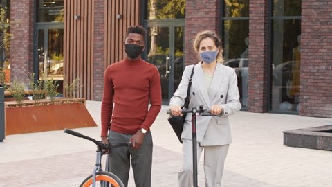 Tilt-up slowmo portrait of young active multi-ethnic business couple with bike and electric scooter wearing smart casualwear and face masks posing for camera standing outdoors in business district