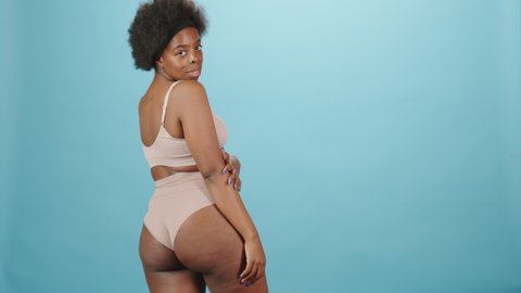 Medium rear-view portrait of beautiful young curvy African-American woman in pink underwear turning to camera standing on isolated blue background