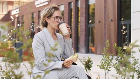 Medium slowmo shot of young Caucasian businesswoman in smart casualwear and eyeglasses on lunch break outdoors, eating sandwich and drinking coffee from plastic cup
