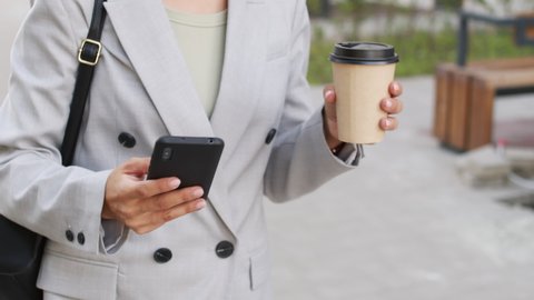 Midsection slowmo shot of unrecognizable elegant businesswoman holding coffee in paper cup and smartphone walking confidently down business district