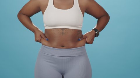 Midsection slowmo shot of unrecognizable plus size African-American woman taking extra fat on sides of her stomach showing it to camera standing in tight sportswear on blue studio background