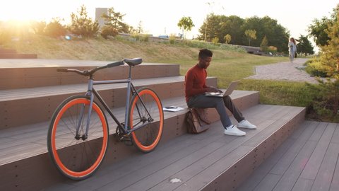 Slowmo stab shot of young African-American businessman or freelancer in smart casualwear working on laptop while sitting on stairs next to his bike outdoors in park at sunset