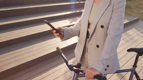 Midsection slowmo shot of unrecognizable businesswoman in pant suit scrolling on smartphone standing with bicycle outdoors on sunny day